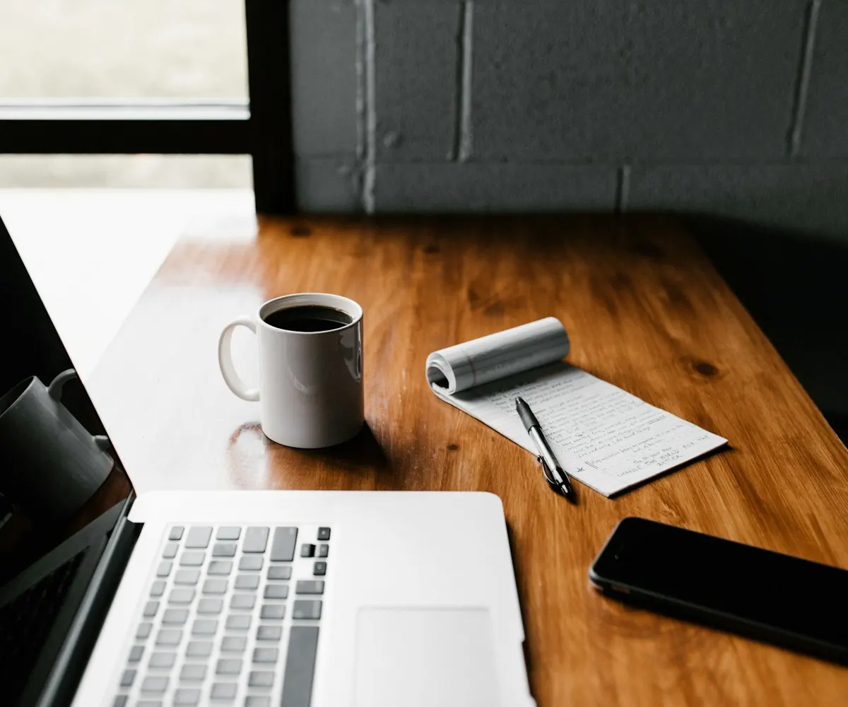 A laptop, notebook and coffee cup arranged on a writing desk.